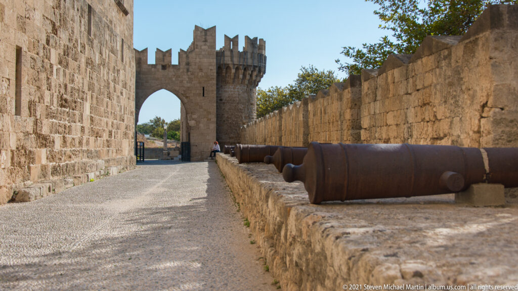 Stone fortification passage leading to a crenellated gateway, with several large iron cannon barrels displayed along the right wall.