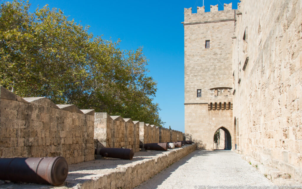 Stone rampart walkway lined with cannon pieces and leading toward a crenellated tower with an arched gateway.