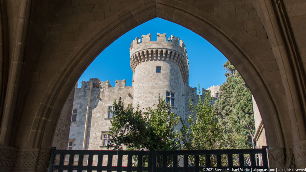 Round crenellated stone tower seen through a pointed arch, with battlements and trees against a bright blue sky.