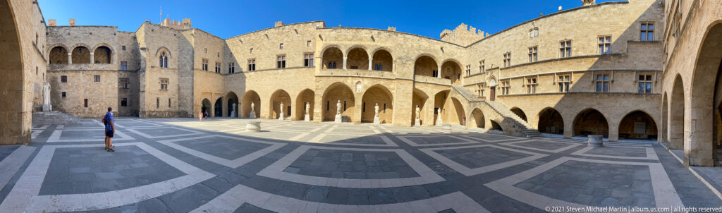Wide palace courtyard with arcades, exterior stairs, upper loggias, geometric paving, and a lone visitor at left.