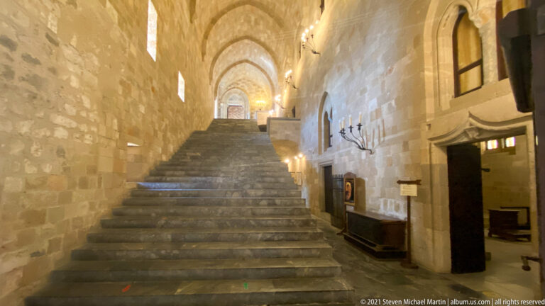Broad worn stone staircase rising through a high vaulted corridor with wall lights and an open doorway at right.