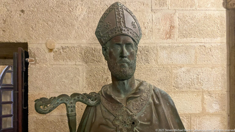 Bronze bust of a bearded ecclesiastical figure in an ornate mitre and vestment, displayed against a pale stone wall.