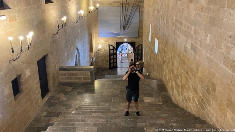 Downward view of a long stone staircase with a visitor, tall masonry walls, wall sconces, and an open doorway below.