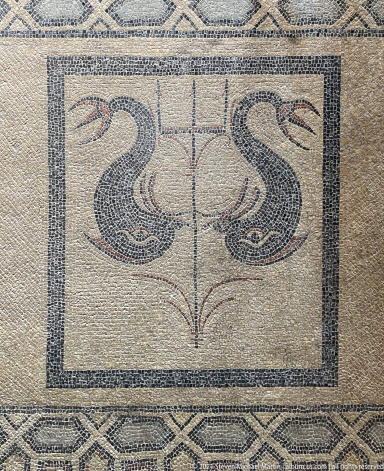Rectangular ancient mosaic panel with paired curving motifs, a central vertical element, and a dark geometric border.