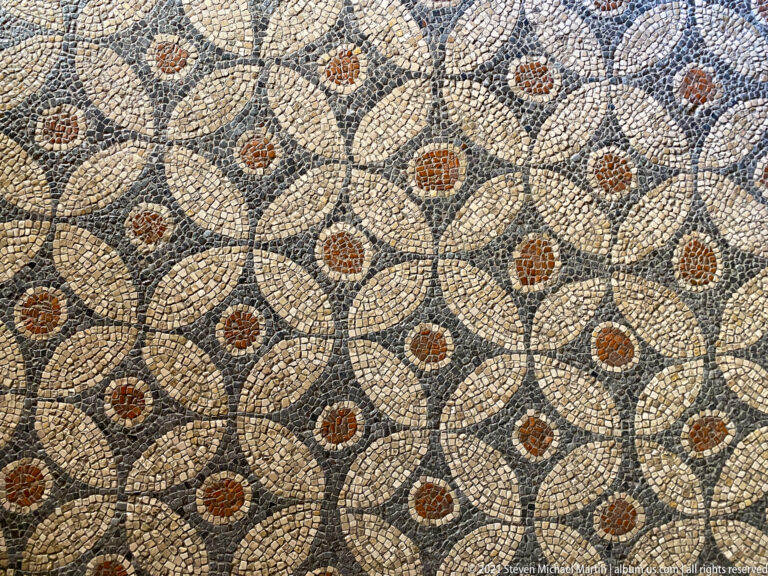 Close view of an ancient mosaic with repeating pale petal shapes and rust-colored rosette centers on a dark ground.