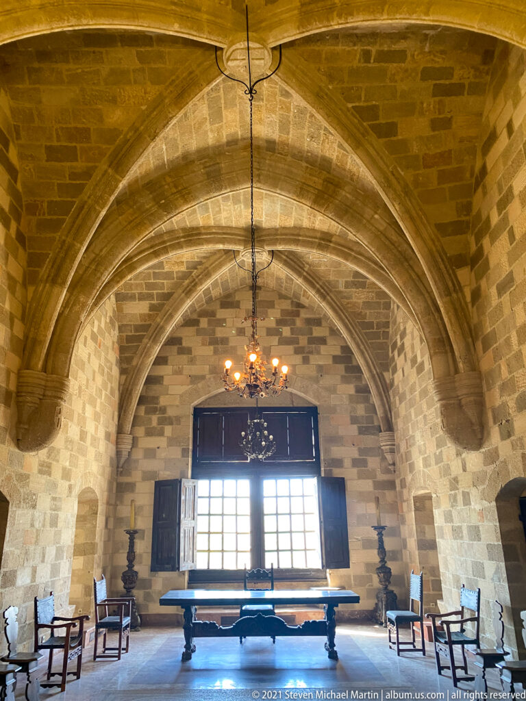 Large vaulted stone hall with a central window, long table, chairs, tall stands, and a hanging chandelier.