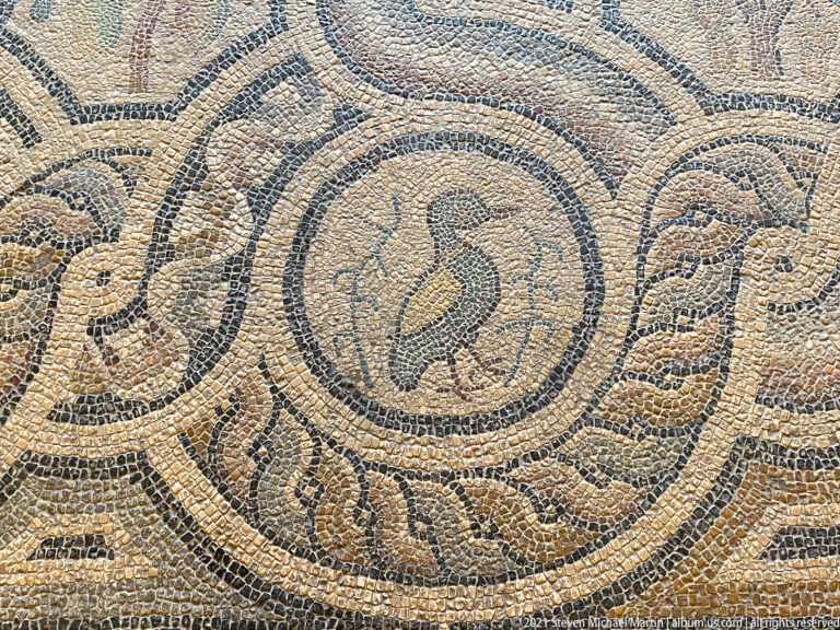 Ancient mosaic with a small bird in a circular medallion surrounded by scrolling geometric and vegetal ornament.