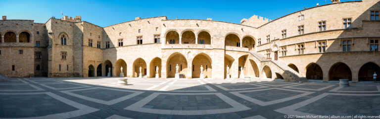 Wide palace courtyard enclosed by stone arcades, with an exterior staircase and large geometric pattern in the paving.