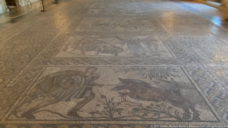Oblique view of a large ancient mosaic floor with multiple rectangular panels containing animals and plant motifs.