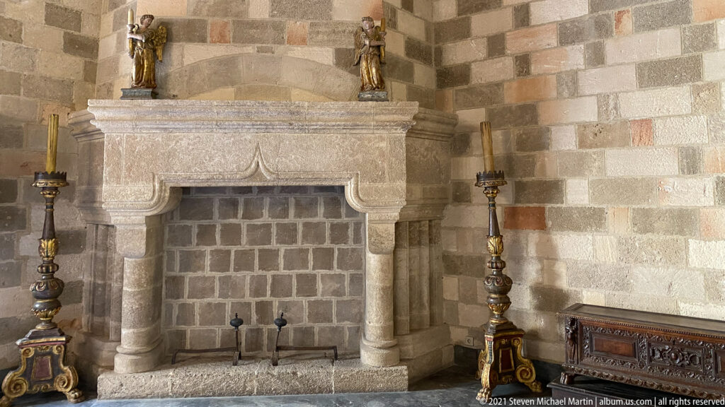 Large carved stone fireplace flanked by tall decorative torchères, with a carved wooden chest against the masonry wall.