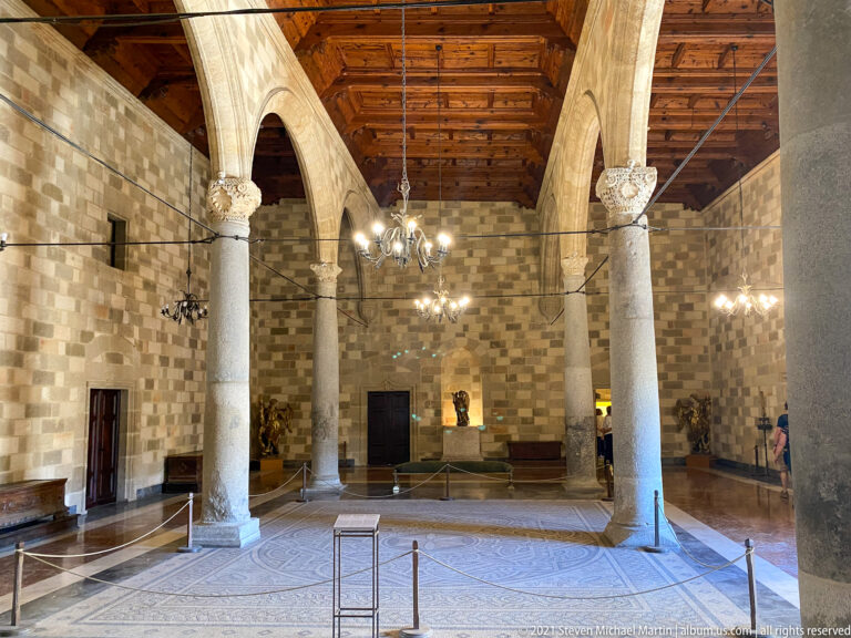 Large palace hall with stone columns, broad arches, chandeliers, a timber ceiling, and historic furnishings around the walls.