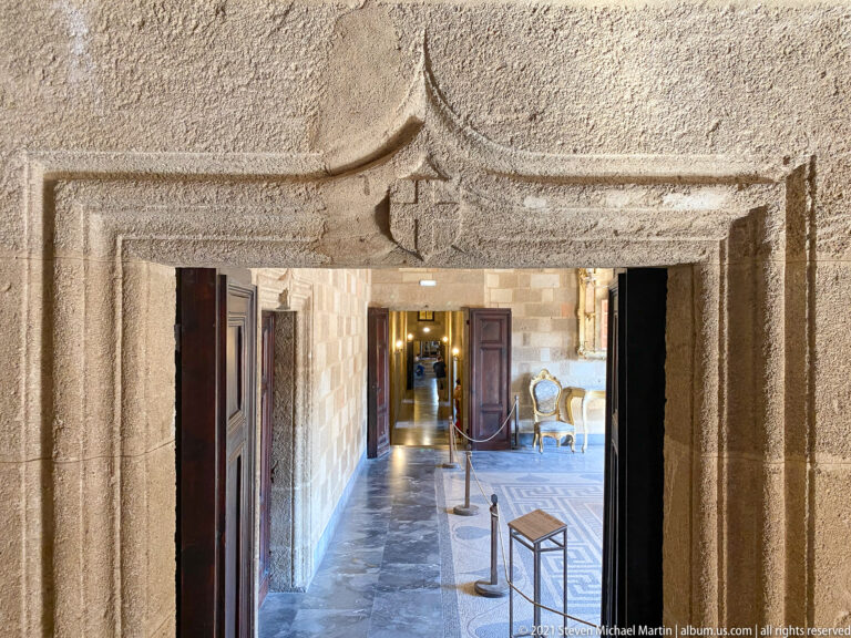 Heavy carved stone doorway framing a sequence of furnished palace rooms with chairs and warm lights beyond.