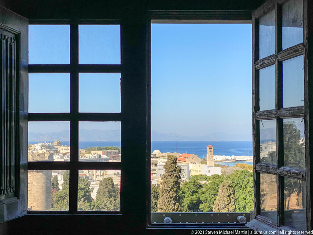 Rhodes town, fortifications, trees, and blue sea seen through a dark open wooden window from inside the palace.