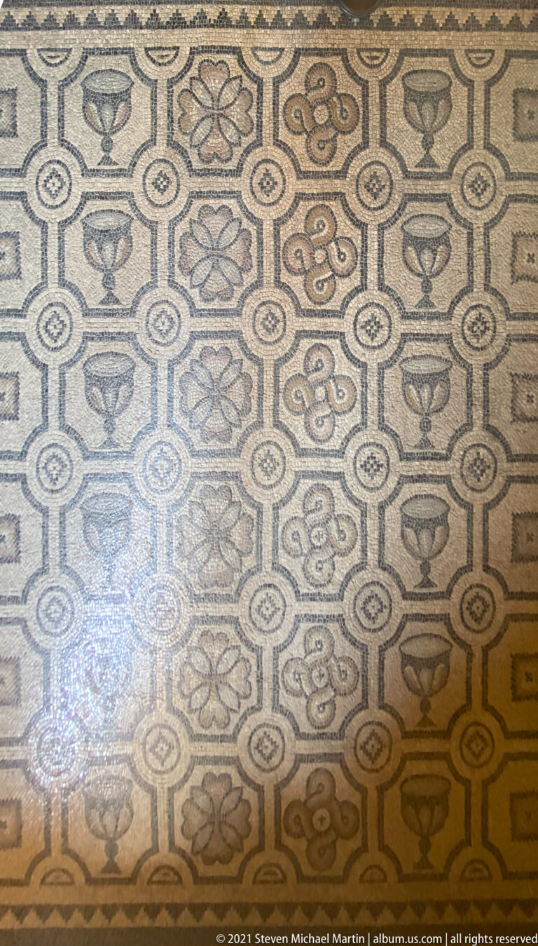Ancient mosaic floor with repeating floral, knot, and vessel-like motifs arranged in linked geometric compartments.