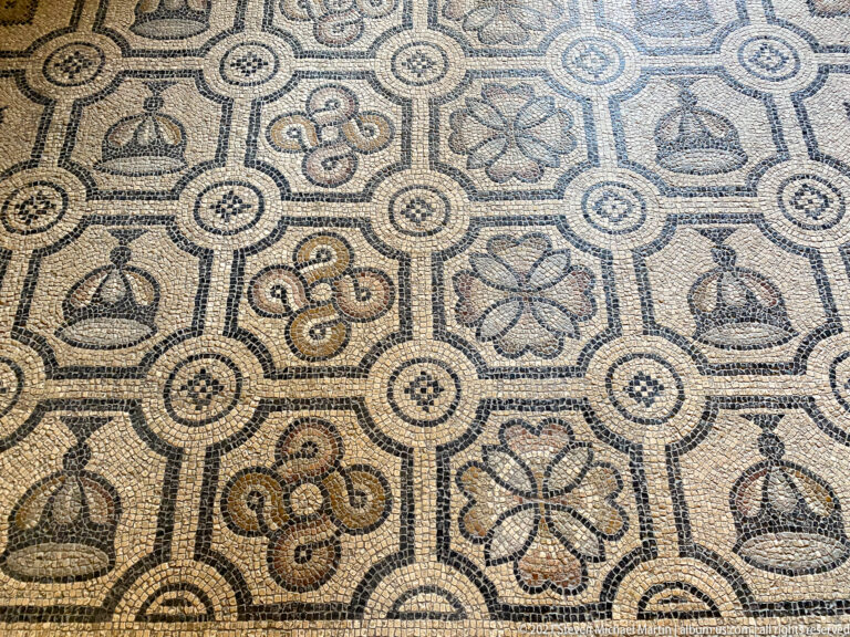 Close mosaic detail with four-petaled flowers, looping knots, and vessel-like motifs linked by circular geometric nodes.