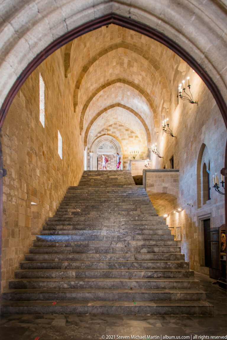 Broad stone staircase rising through a long barrel-vaulted hall lit by small windows and wall lamps.