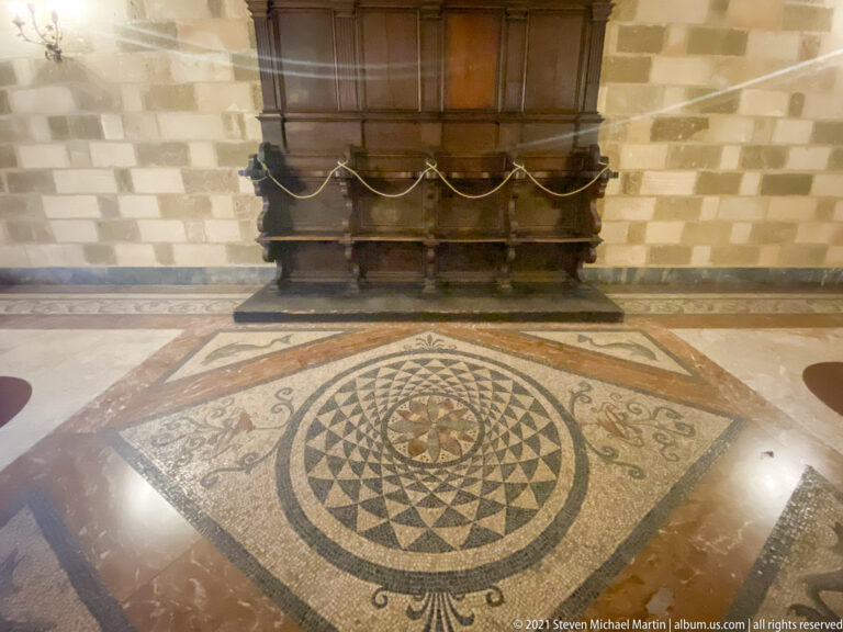 Circular geometric mosaic medallion in a stone floor directly before a large dark wooden chest against the wall.