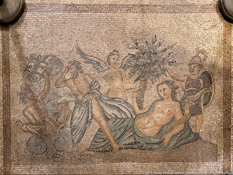 Ancient rectangular mosaic with several classical figures, including a reclining figure at right and standing figures around the center.