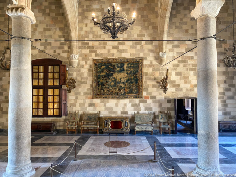 Columned palace hall with a wall tapestry, historic chairs, chandelier, timber ceiling, and patterned stone floor.
