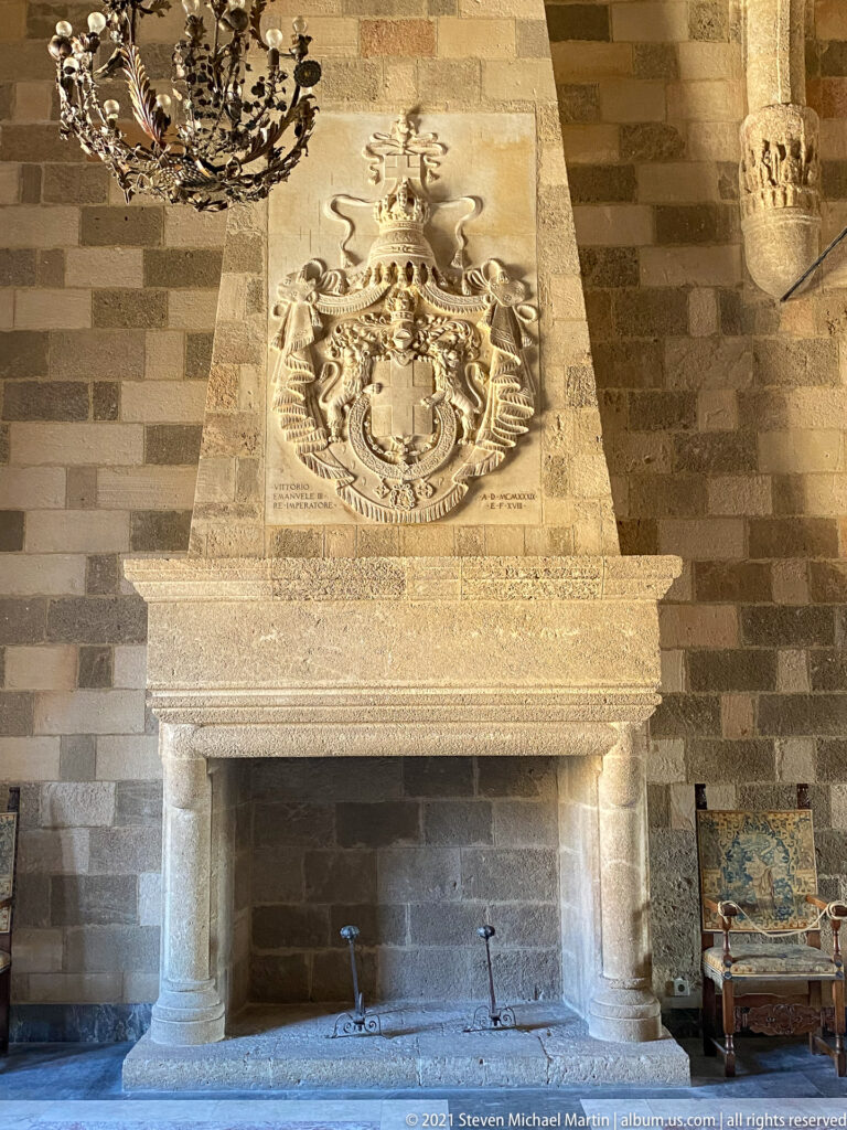 Large stone fireplace beneath an elaborate carved coat of arms, set against banded masonry in a palace room.