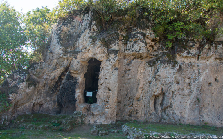 Rock-cut doorway in a weathered limestone cliff, with low stone remains and vegetation at the School of Aristotle site.