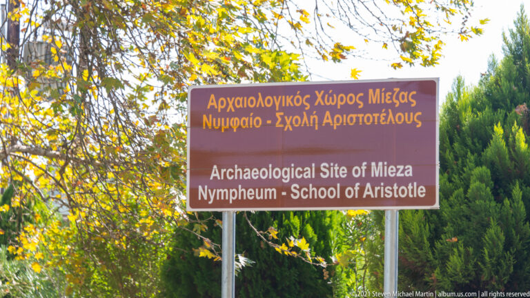 Brown bilingual sign reading “Archaeological Site of Mieza, Nymphaeum – School of Aristotle,” surrounded by trees.