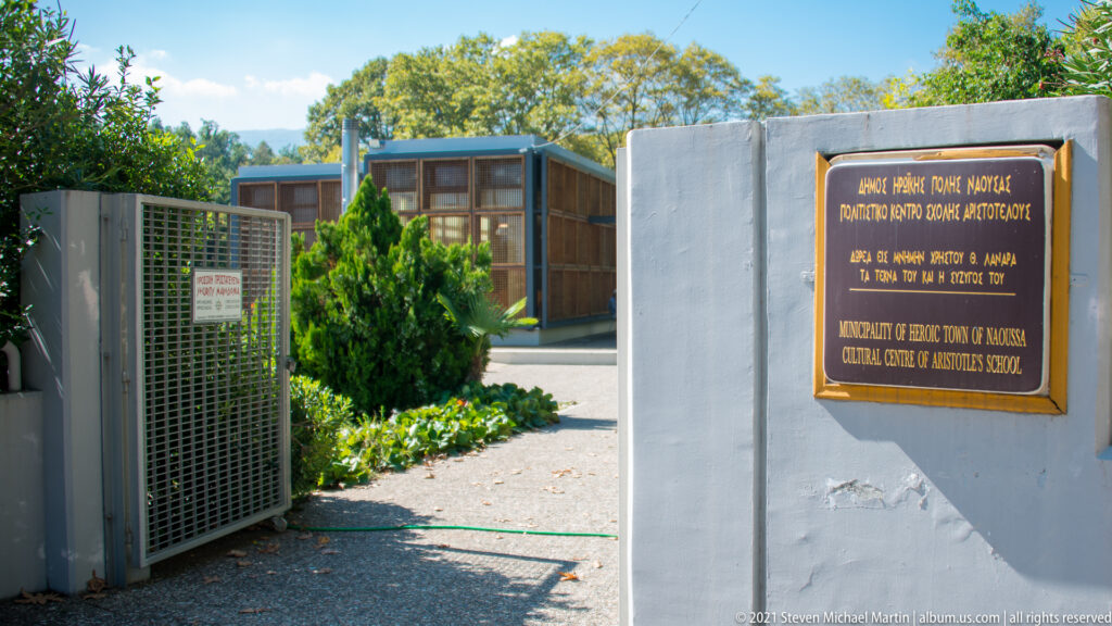 Modern entrance with a dark plaque referring to an Aristotle cultural center, landscaped shrubs, and contemporary buildings beyond.