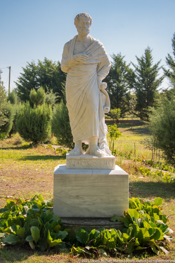 White marble statue of a bearded, draped Aristotle standing on a rectangular pedestal in a landscaped outdoor setting.