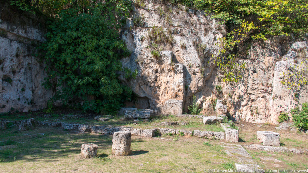 Grassy archaeological court with low foundations and stone blocks below a vegetation-covered, rock-cut limestone wall at Mieza.