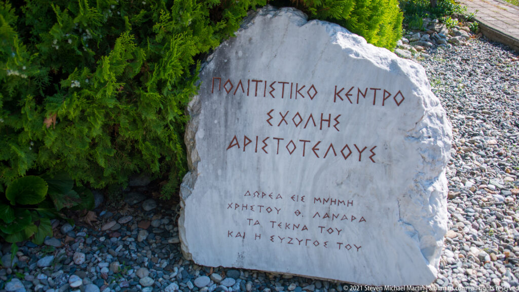Pale irregular stone marker with red Greek lettering naming the School of Aristotle cultural center, set in gravel beside greenery.
