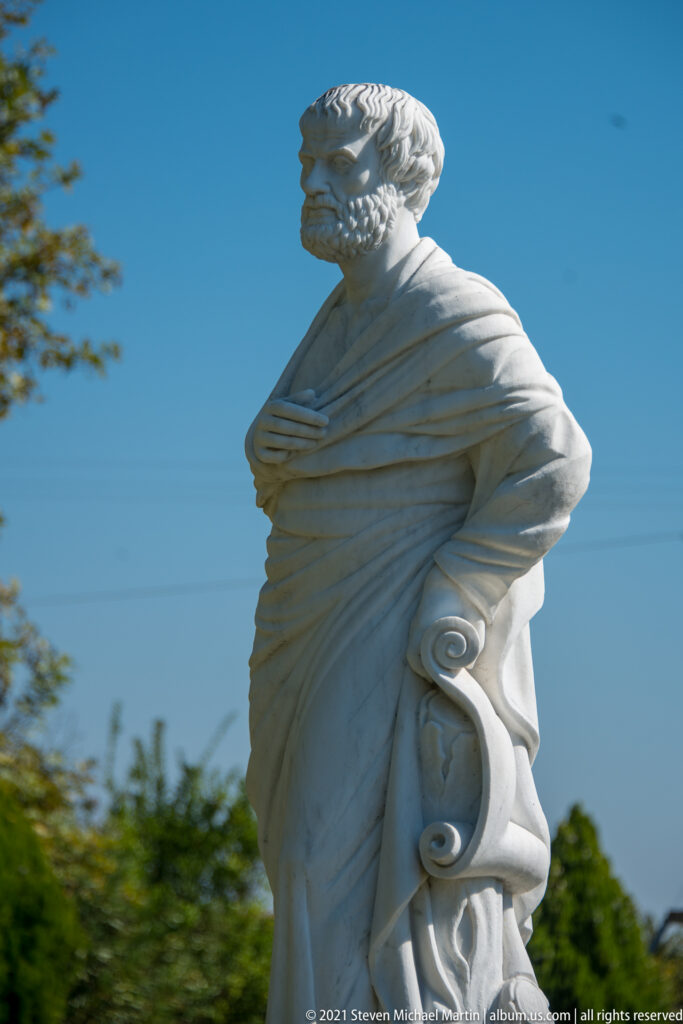 Low-angle three-quarter view of a white marble Aristotle statue with curled beard, heavy drapery, and rolled forms at the side.