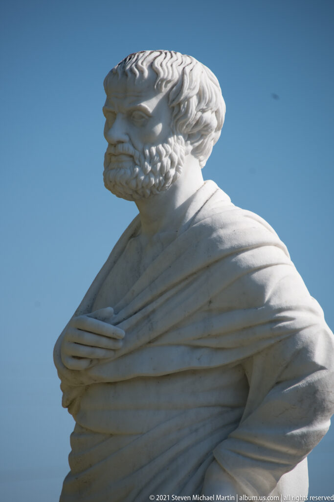 Close view of the white marble Aristotle statue’s bearded head, shoulders, and deeply folded cloak against blue sky.