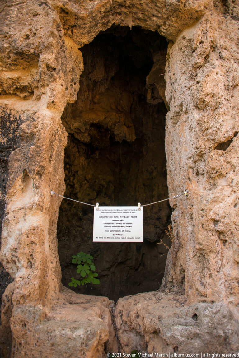 Close view of a deep rock-cut recess with rough stone edges, a protective cord, interpretive sign, and small plants.