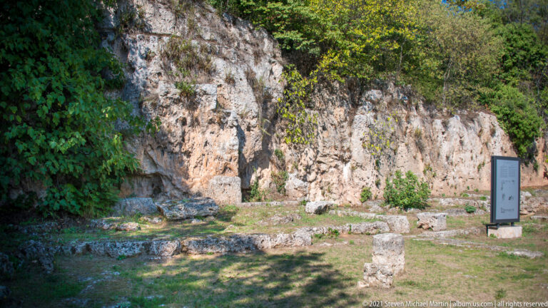 Oblique view of low stone foundations beside a long rock-cut cliff, with vegetation and an information panel at right.