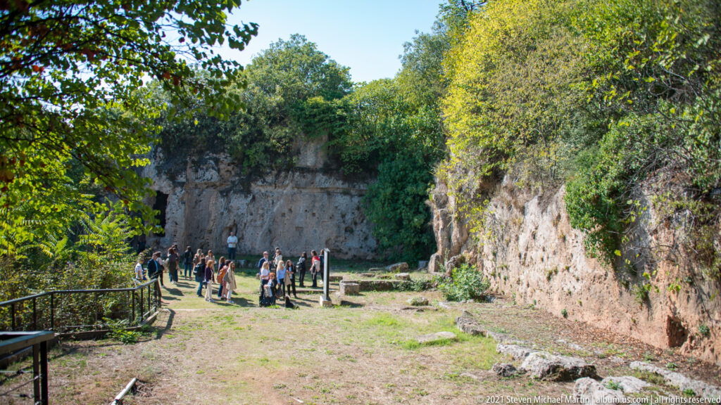 Visitors gathered on a grassy path between rock-cut cliffs and low archaeological remains at the Mieza site.