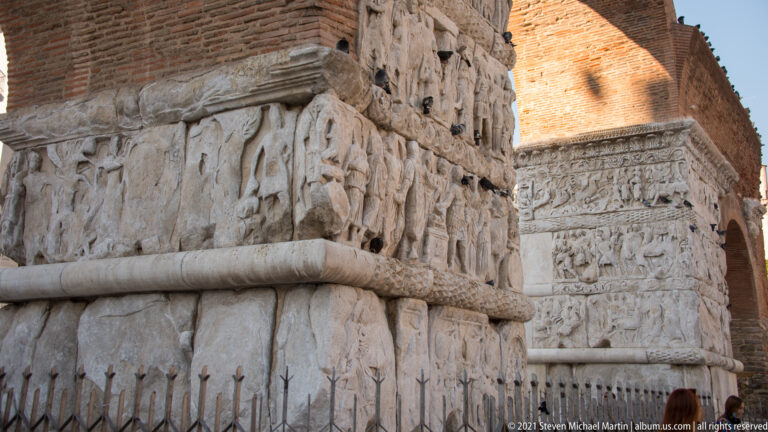 Carved marble relief registers on a pier of the Arch of Galerius beneath a Roman brick arch.