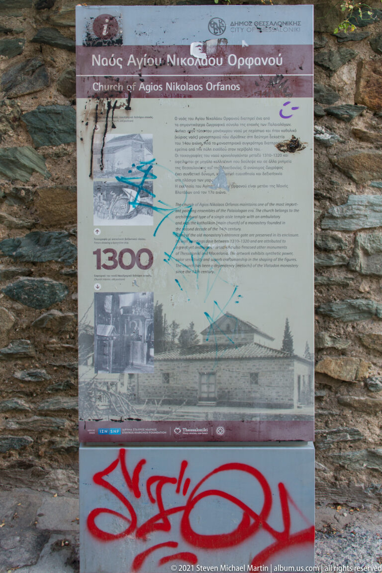 Graffiti-marked interpretive panel for Agios Nikolaos Orfanos mounted against a stone wall.