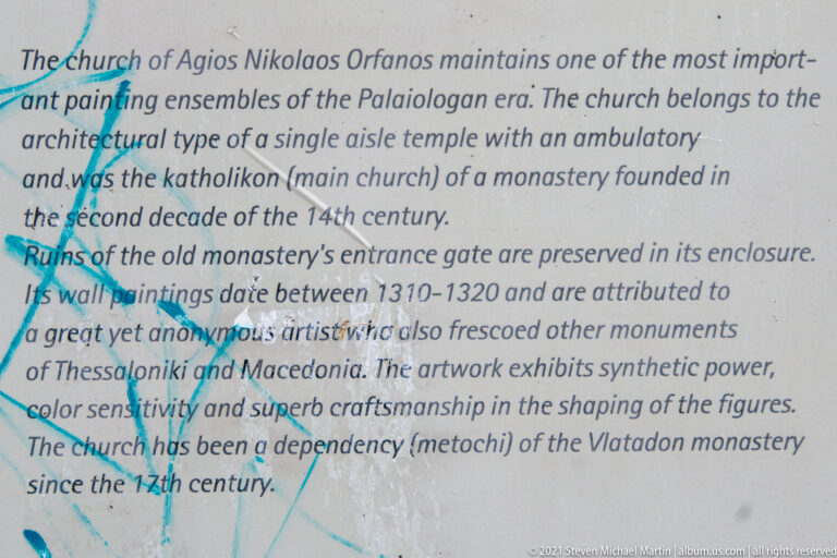 Close view of English interpretive text about Agios Nikolaos Orfanos, crossed by blue graffiti.