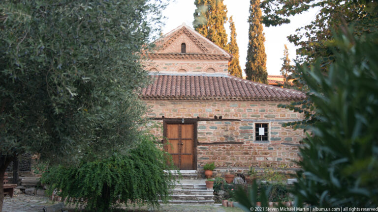 Brick-and-stone exterior of Agios Nikolaos Orfanos framed by trees and garden vegetation.
