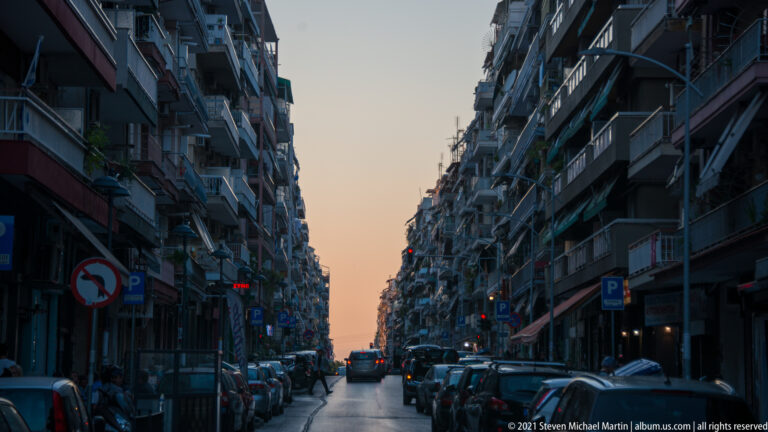 Narrow city street lined with parked cars and apartment balconies at dusk.