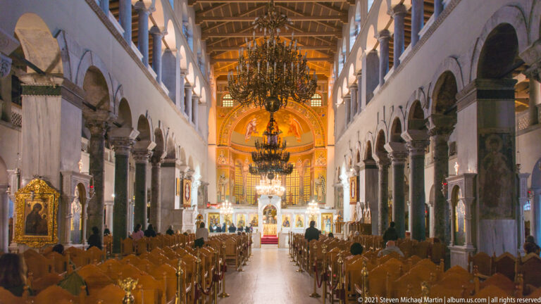 Long basilican nave with marble columns, arches, chandeliers, wooden seating, and illuminated sanctuary.