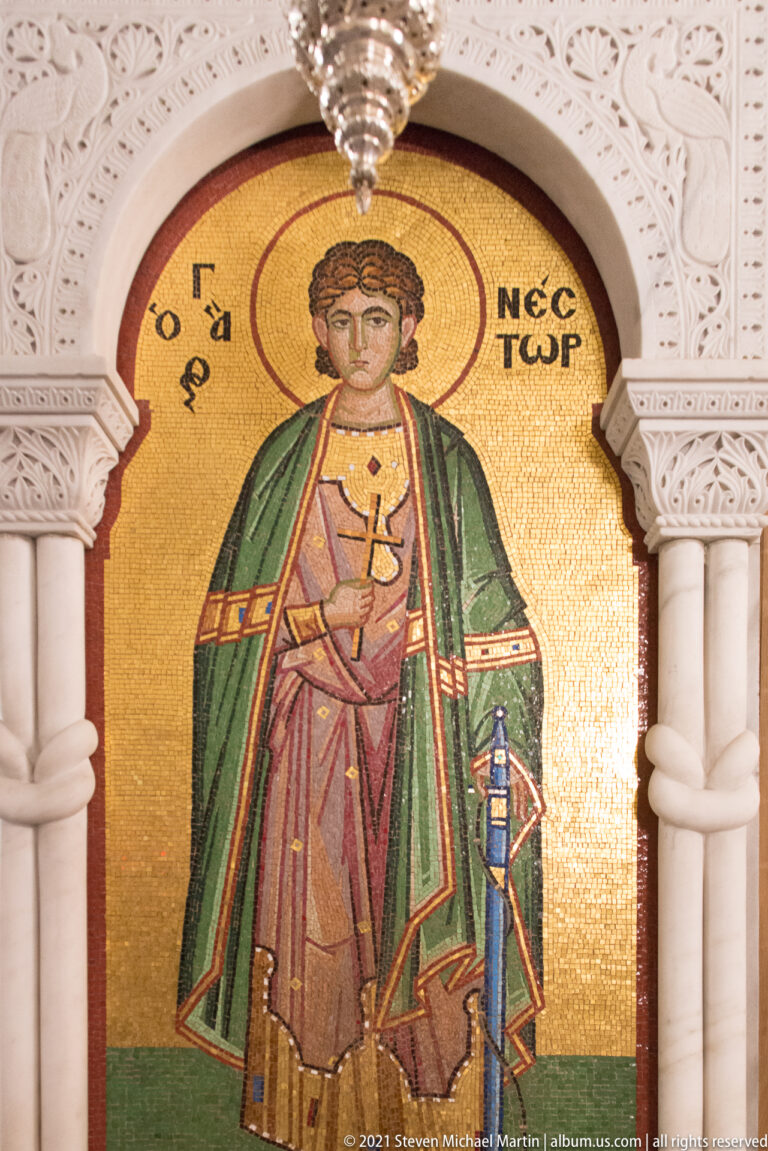 Gold-ground mosaic of Saint Nestor standing with a cross in an arched white frame.