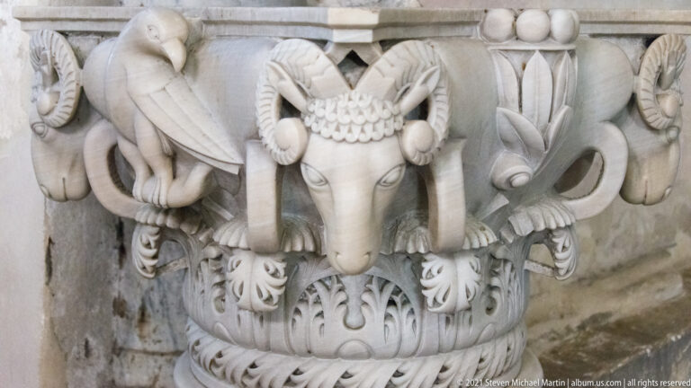 Close view of a carved marble capital with a projecting ram’s head and curled horns.