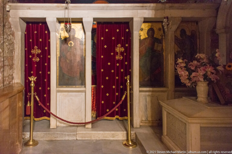 Red cross-embroidered curtains within a marble-framed shrine area, flanked by painted saints.