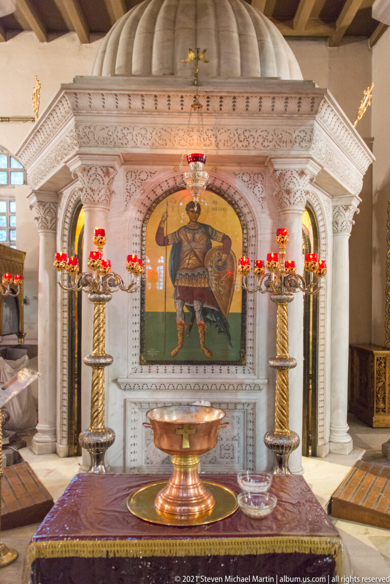 White marble shrine with saint icon, red lamps, candle stands, and bronze basin.