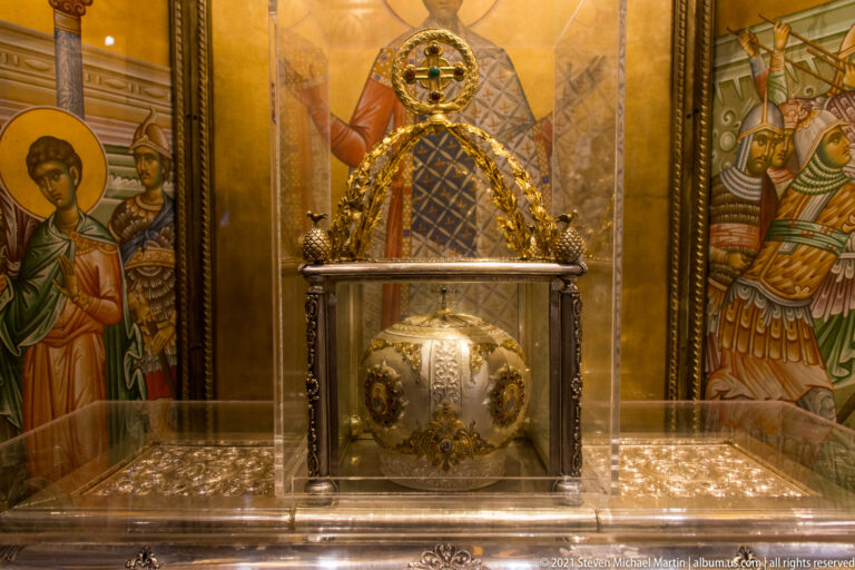Ornate silver reliquary displayed behind glass amid gilded metalwork and painted saints.