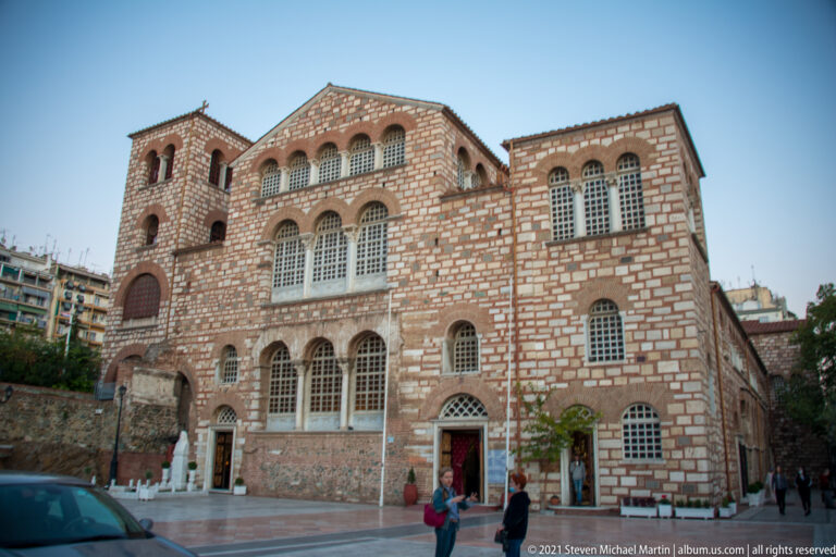 Brick-and-stone Byzantine church façade with arched windows and a bell tower beside a Thessaloniki street.