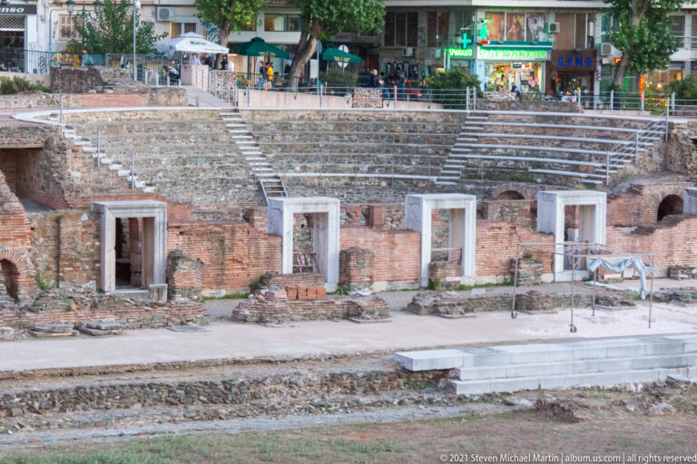 Close view of ancient stepped seating, brick doorways, and modern businesses above the archaeological site.