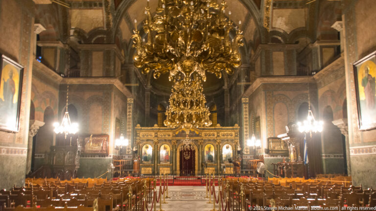 Symmetrical nave view with gilded chandelier, wooden seating, and iconostasis at the far end.