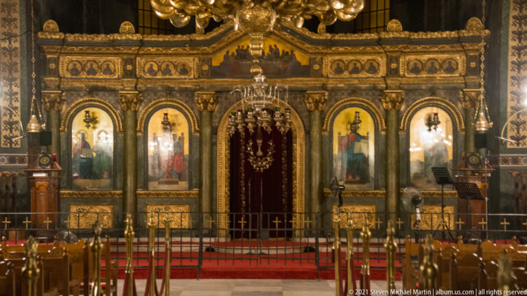 Ornate gilded iconostasis with saint icons, red central curtain, lamps, and sanctuary rail.